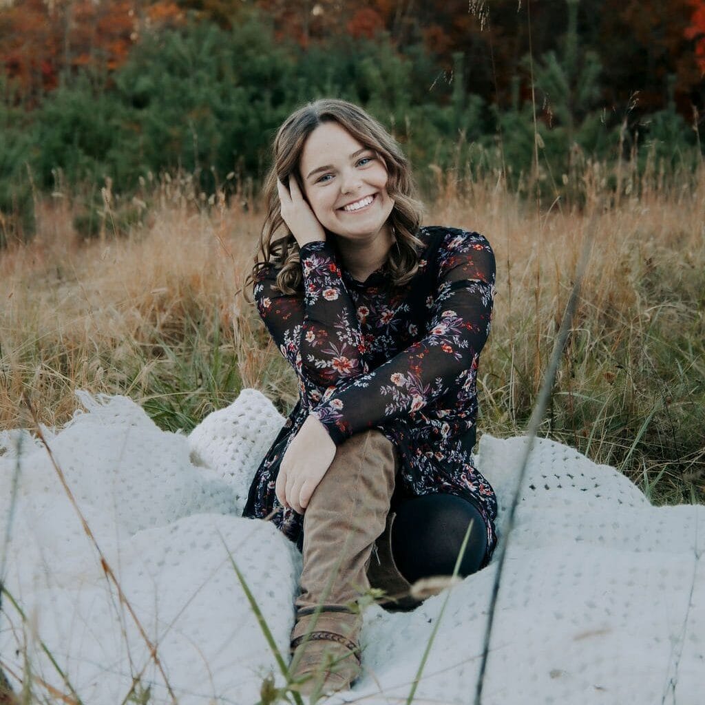 allyson sits on a blanket in a grassy field, smiling at the camera with hand in her hair.
